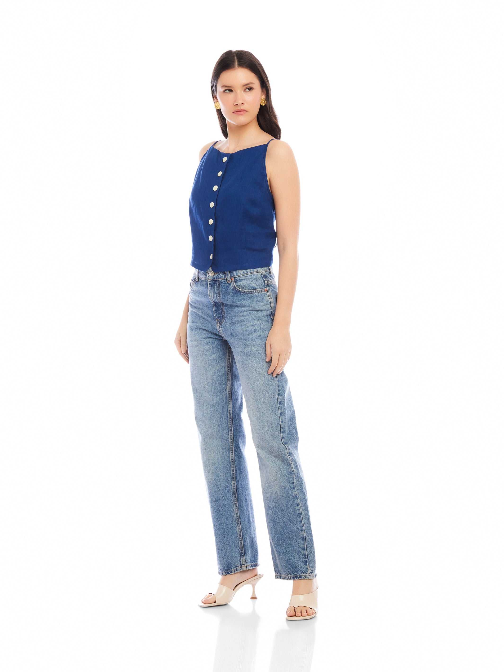 Woman wearing Elis sleeveless button-up top in blue linen with light blue jeans and heels