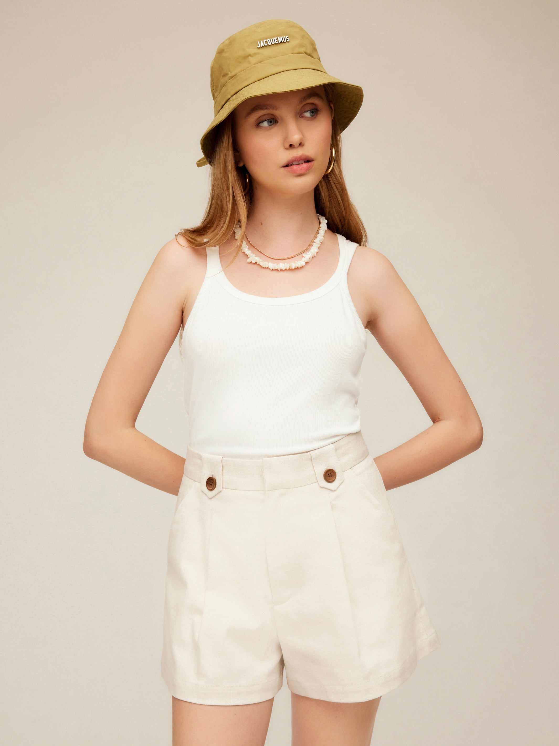 Woman wearing Cassidy Pleated Shorts in beige with a white tank top and beige bucket hat.