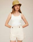 Woman wearing Cassidy Pleated Shorts in beige with a white tank top and beige bucket hat.