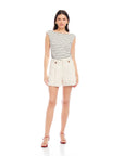 Woman wearing Cassidy Pleated Shorts in light beige with front pleats and button detail, paired with a striped sleeveless top and red sandals