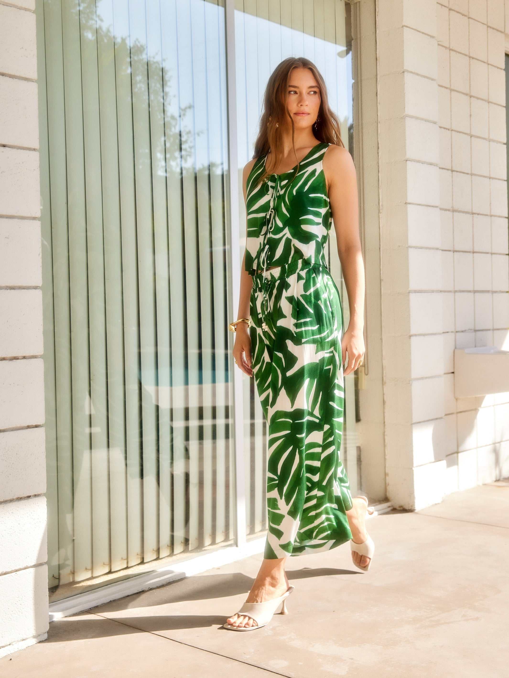 Woman wearing green and white Monstera print Etta Cropped Drawstring Pants with wide leg and sleeveless top standing outdoors by glass doors