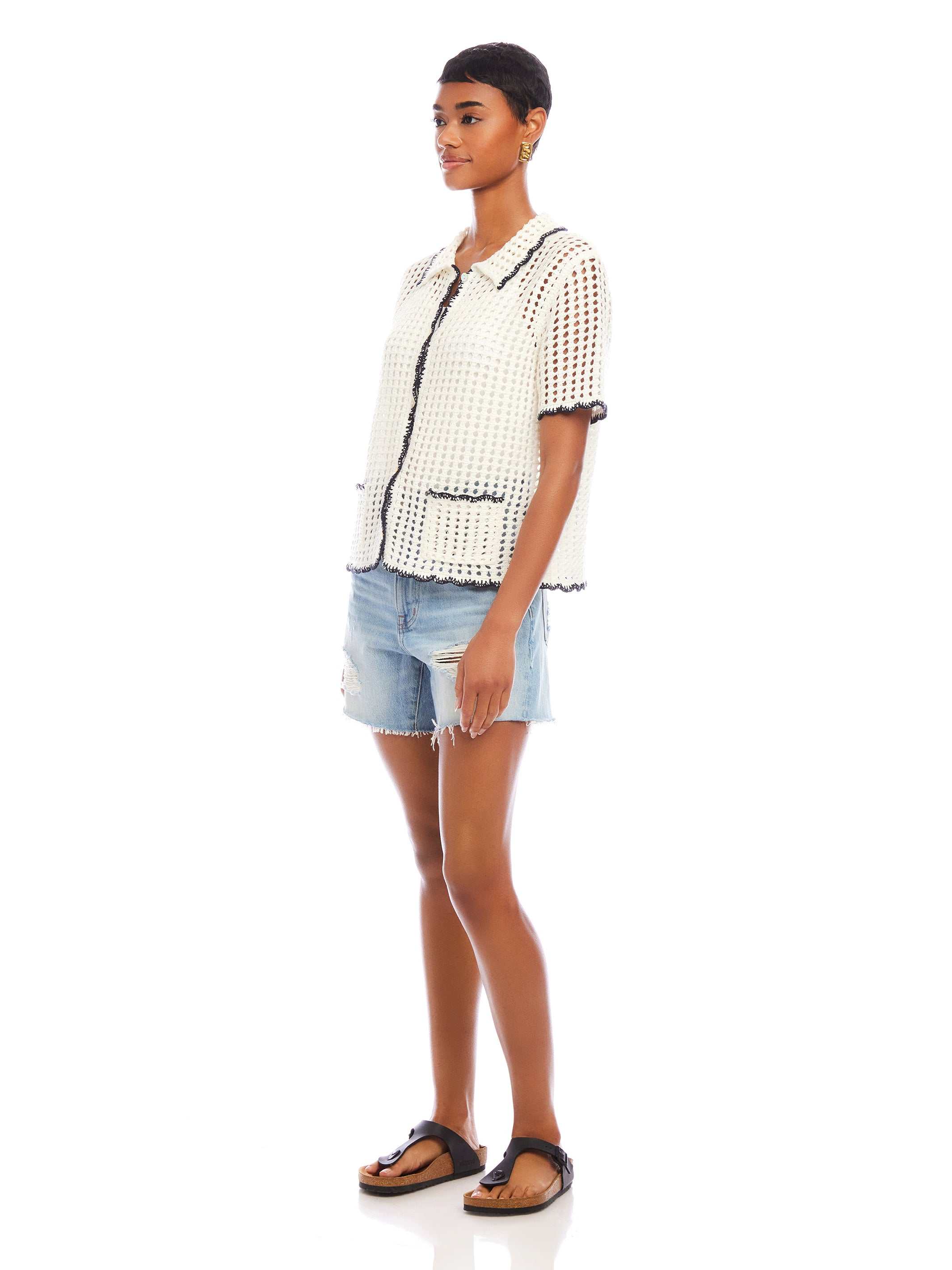 Woman wearing a white Gemma Crochet Shirt with contrasting black trim, boxy silhouette, short sleeves, front pockets, and scalloped edges.