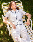 Woman wearing white Gemma Crochet Shirt with contrast black stitching, short sleeves, scalloped edges, and two front pockets while lounging on a striped chair outdoors