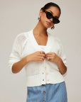 Woman wearing white Ellen Crochet Cardigan with V-neck, ribbed edges, and long sleeves, styled with sunglasses and blue skirt.