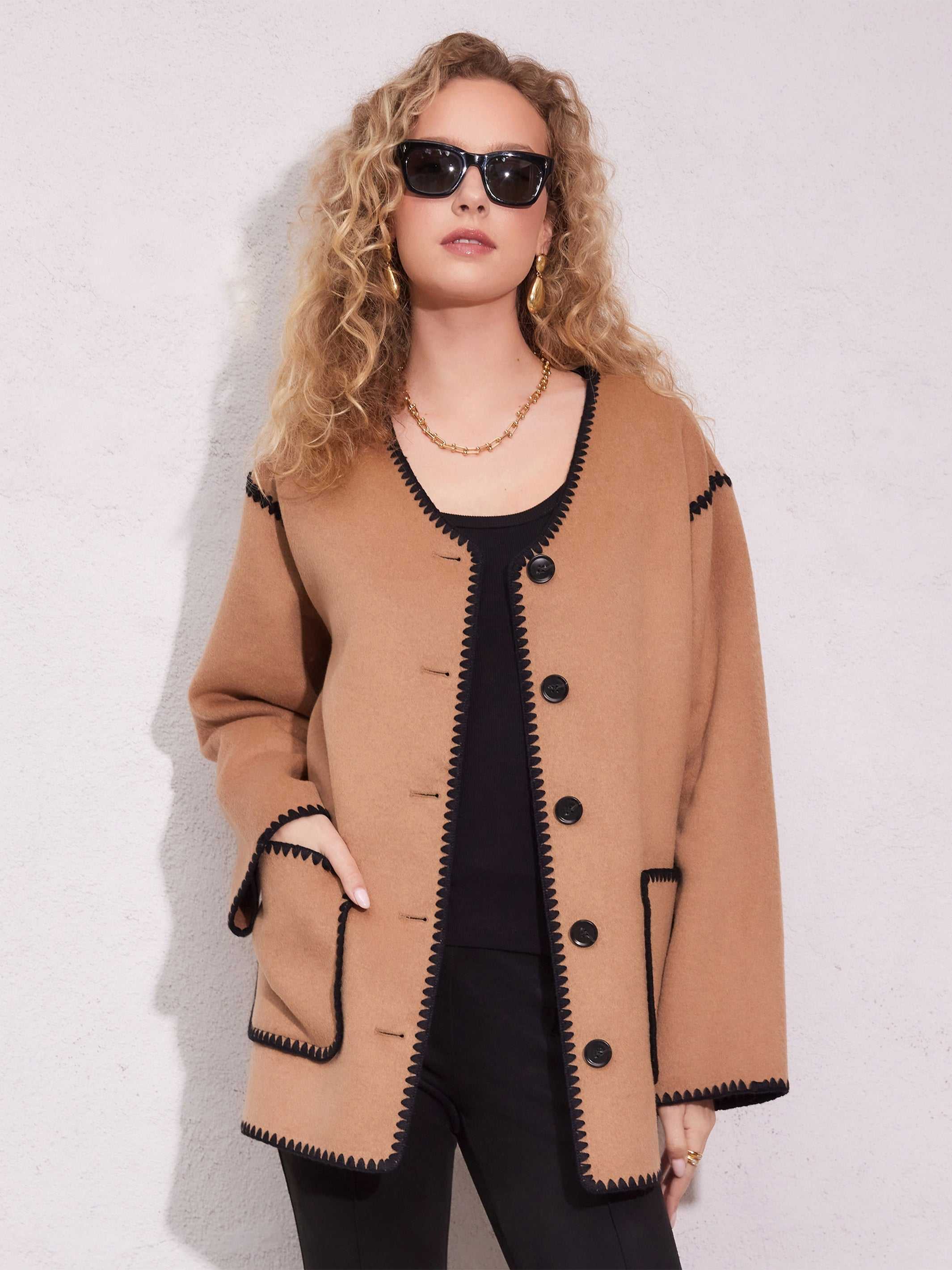 Woman wearing tan Nico Jacket with black trim, black buttons, long sleeves, and V-neck over black top and pants, accessorized with sunglasses and gold jewelry standing against a light wall