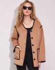 Woman wearing tan Nico Jacket with black trim, black buttons, long sleeves, and V-neck over black top and pants, accessorized with sunglasses and gold jewelry standing against a light wall