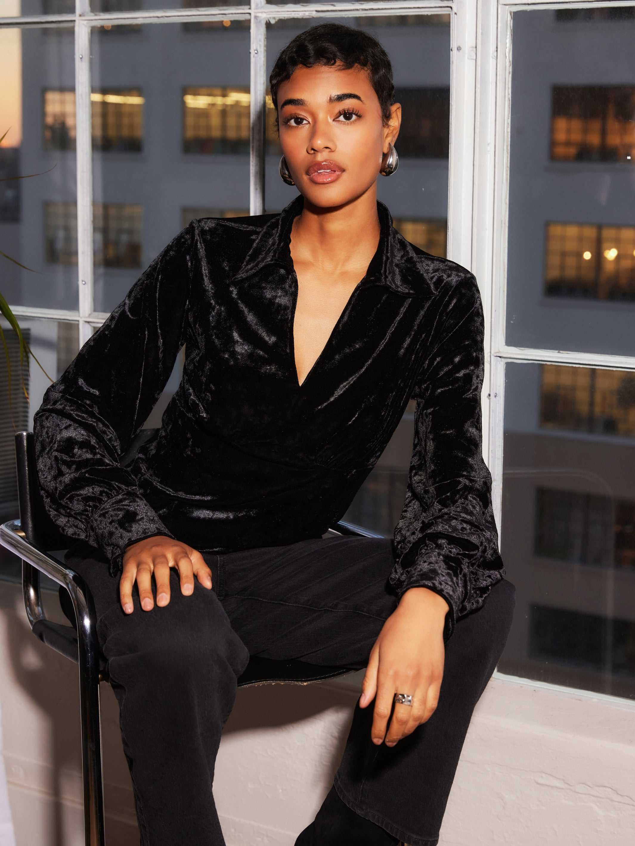 Woman wearing black Bianca Collared Shirred Top in crushed velvet with long sleeves and button cuffs sitting indoors by window