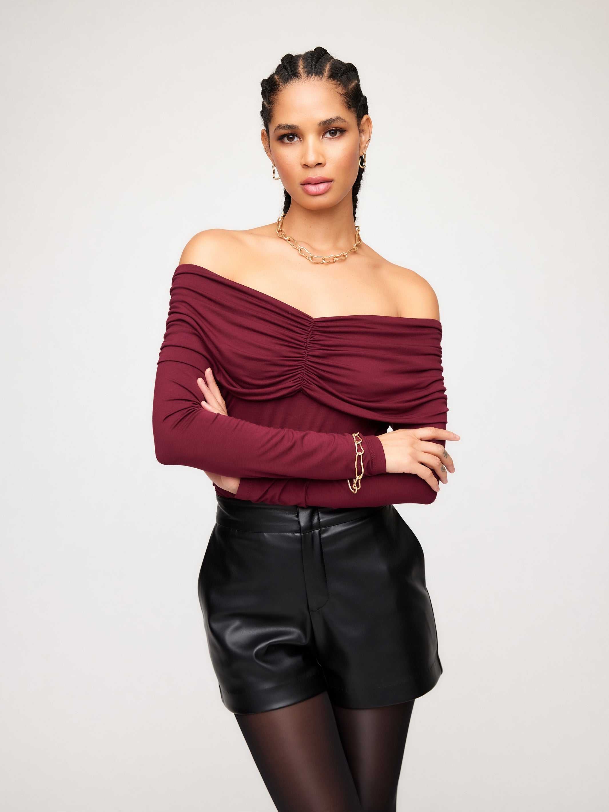 Woman wearing a burgundy Bella Off-The-Shoulder Top with long sleeves and shirring detail paired with black leather shorts
