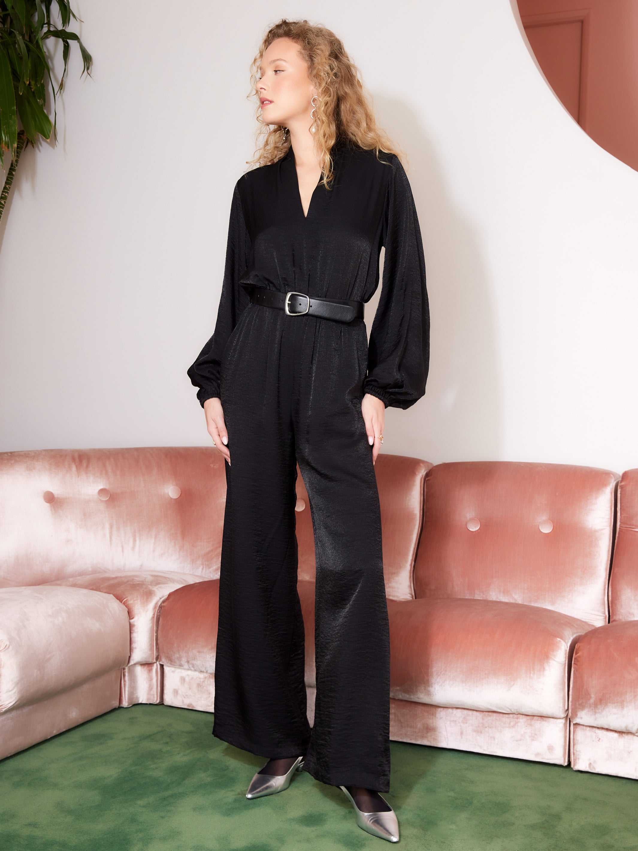 Woman wearing black Skyler Jumpsuit with long blouson sleeves, V-neck, and belted waist standing indoors