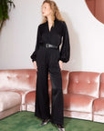 Woman wearing black Skyler Jumpsuit with long blouson sleeves, V-neck, and belted waist standing indoors