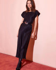 Woman wearing black silky Emmeline Bias Dress with cap sleeves and round neck standing against pink wall