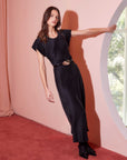 Woman wearing black silky Emmeline Bias Dress with cap sleeves and fitted waist standing indoors near circular window