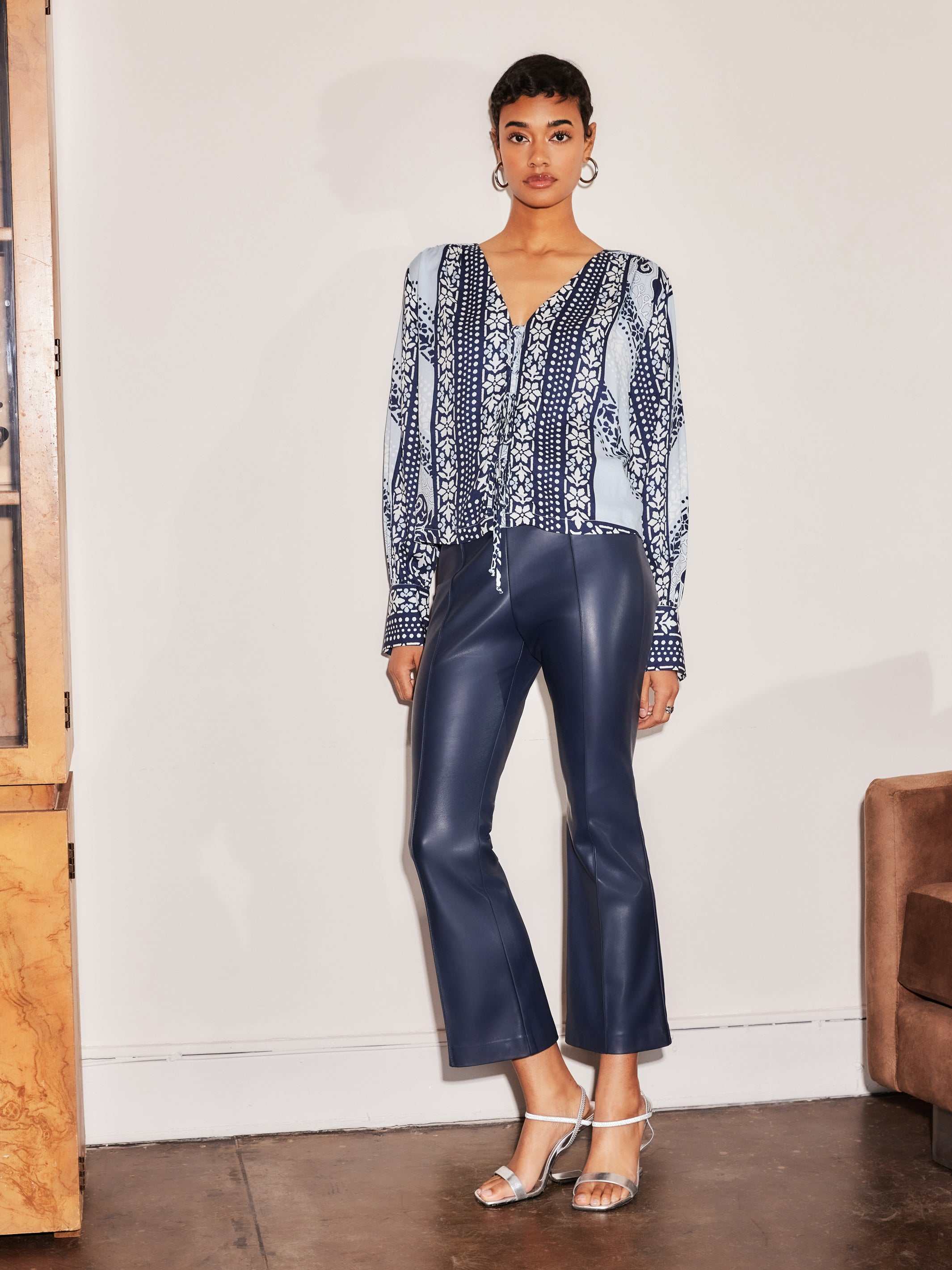 Woman wearing navy Sigourney Pintuck Pants made of faux leather with flared cropped legs and front pintuck seams.