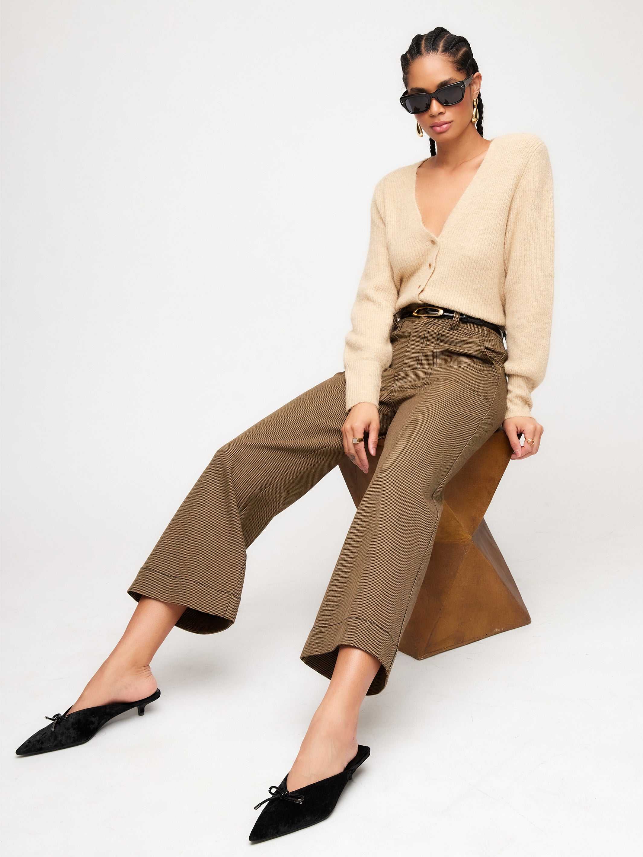 Woman wearing beige Kennedy Cardigan with bishop sleeves, brown wide-leg pants, black pointed-toe flats, and sunglasses sitting on a geometric stool
