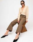 Woman wearing beige Kennedy Cardigan with bishop sleeves, brown wide-leg pants, black pointed-toe flats, and sunglasses sitting on a geometric stool