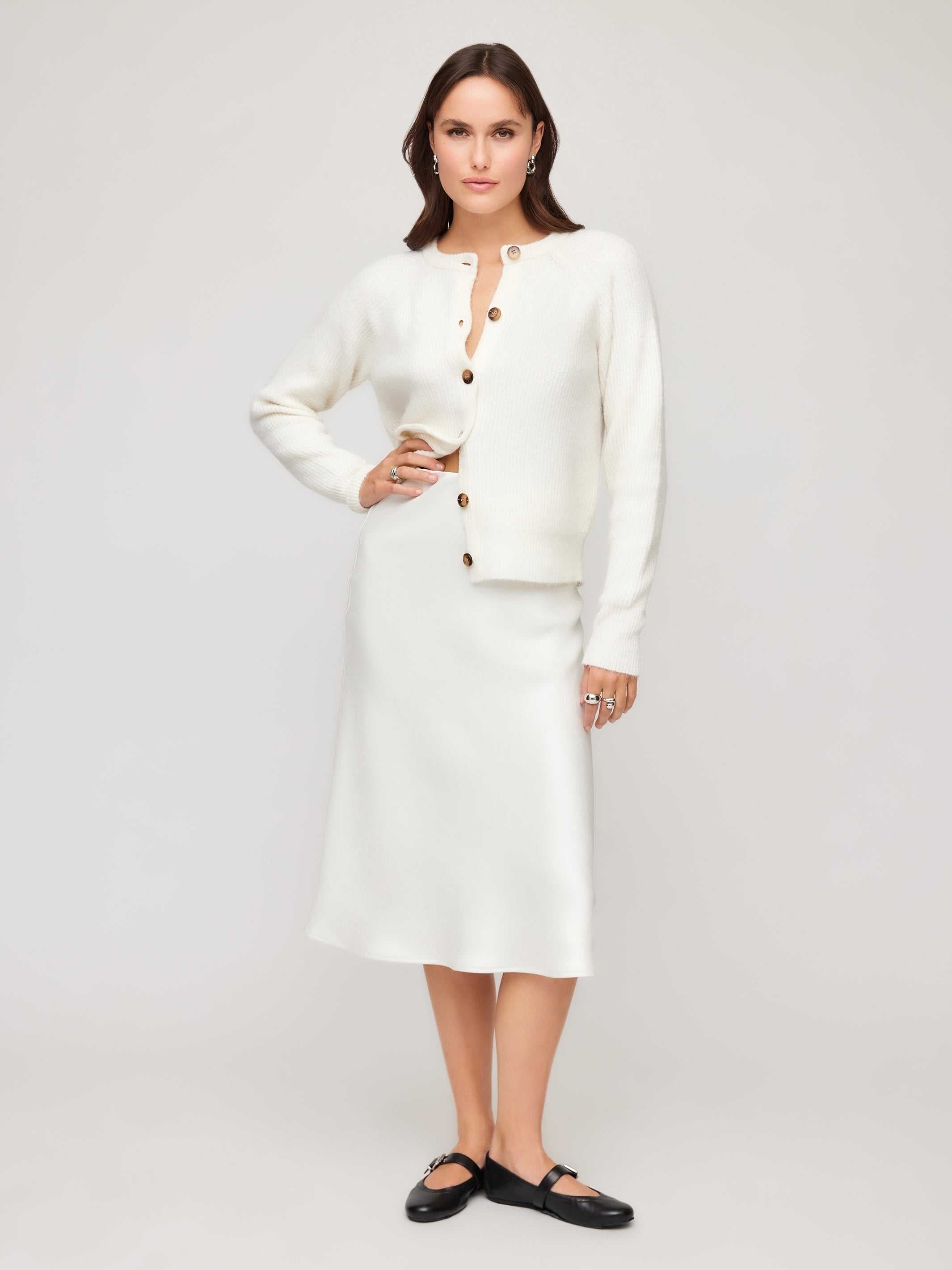Woman wearing a white satin Mia Bias Skirt with an ivory button-up cardigan and black flats standing against a neutral background.