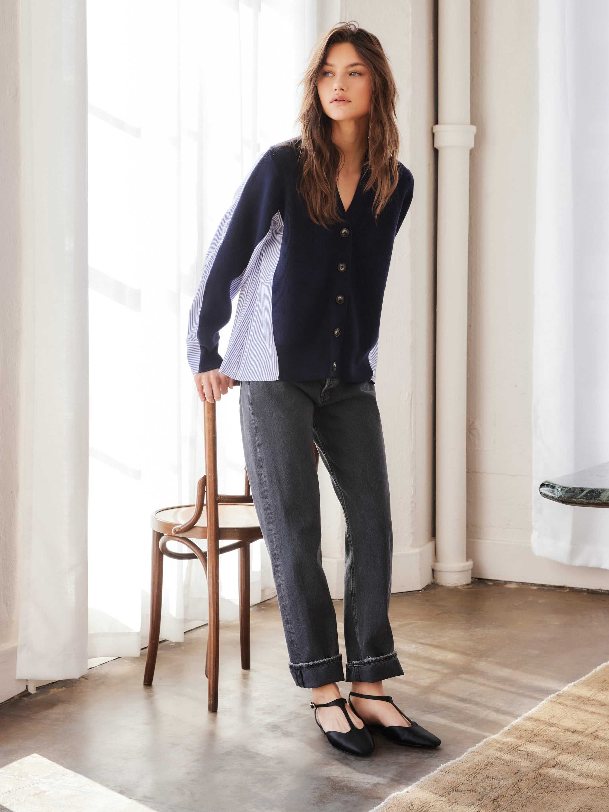Woman wearing navy Celine Mixed Media cardigan with white and blue stripe panels, paired with black cuffed pants and black flats, standing indoors next to a wooden chair