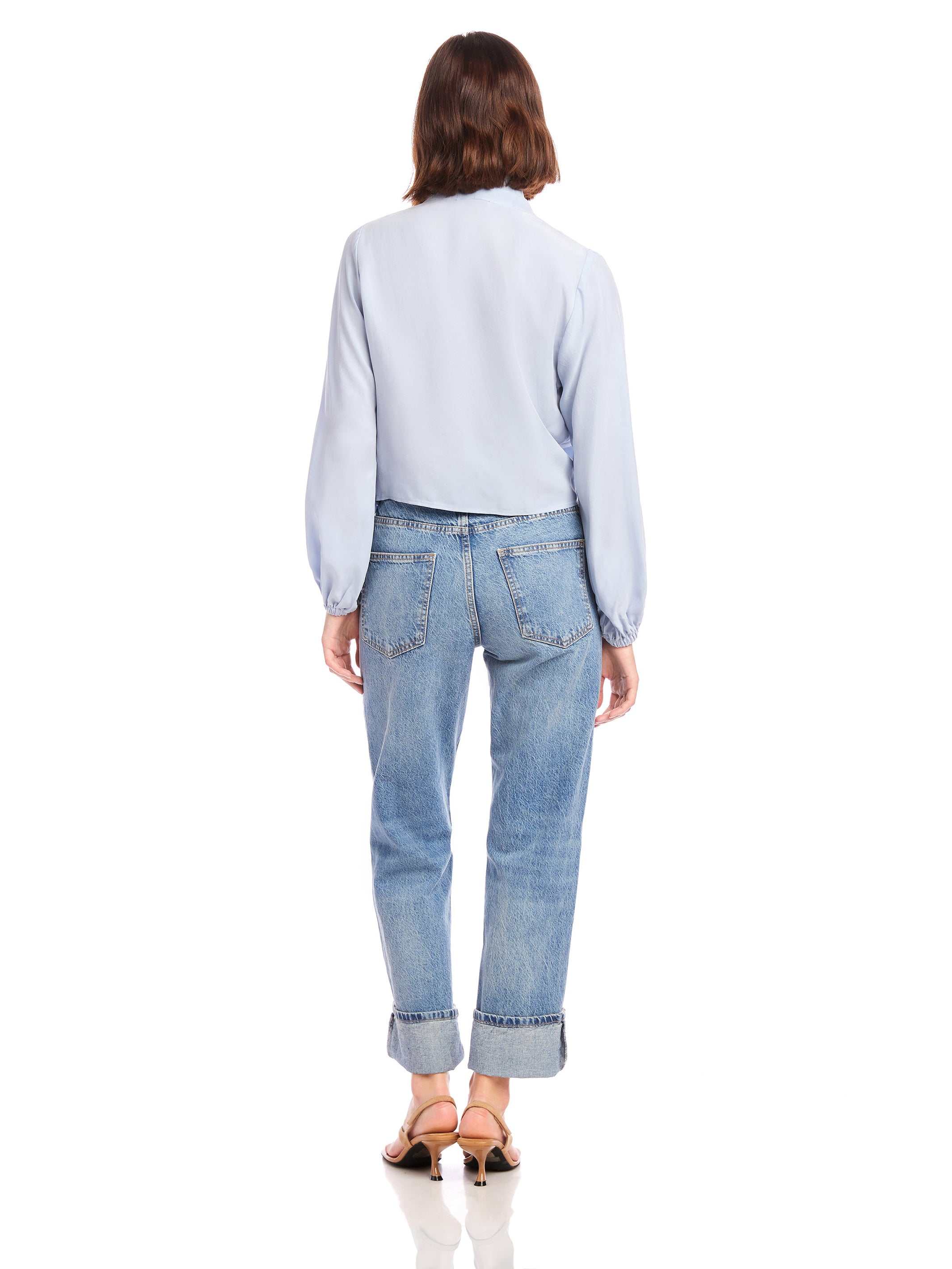 Woman wearing a light blue Capri Tie-Front Blouse with long blouson sleeves, elasticized cuffs, paired with cuffed blue jeans and sandals, shown from the back.
