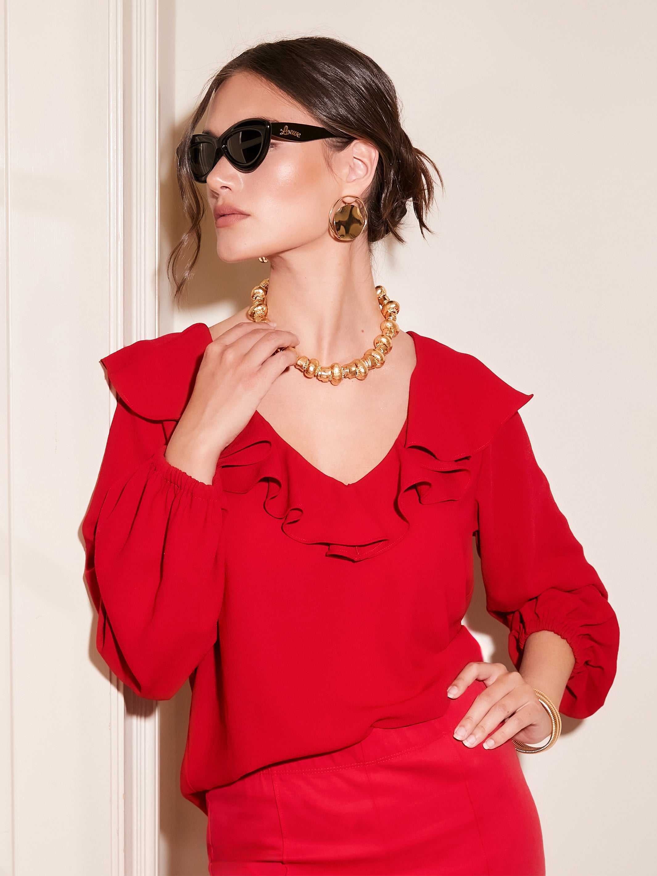Woman wearing red Rita Ruffle Blouse with V-neck and front ruffle detail paired with gold accessories and sunglasses