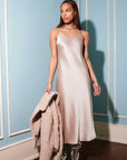 Woman wearing a silky satin Bias Midi Dress with adjustable spaghetti straps and V-neck holding a beige coat indoors