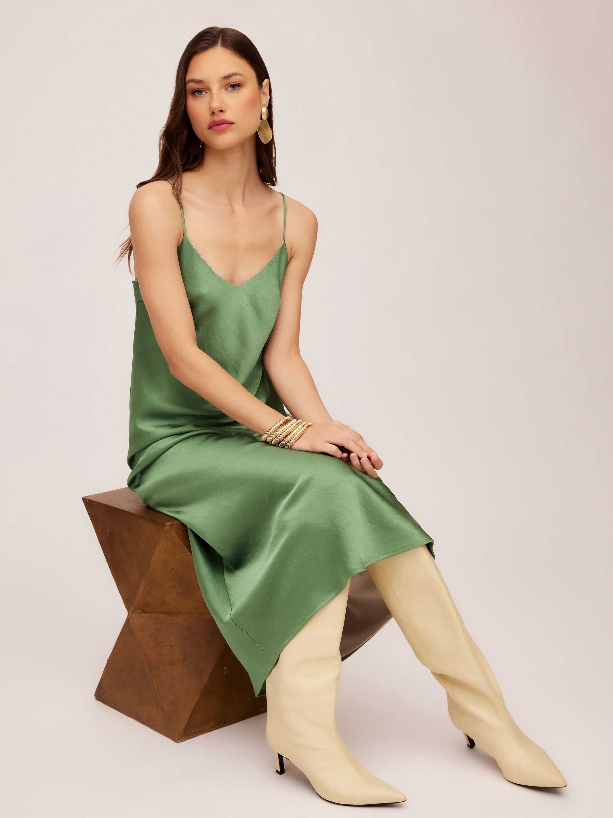 Woman wearing green bias midi dress with adjustable spaghetti straps and V-neck sitting on geometric wooden stool
