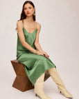 Woman wearing green bias midi dress with adjustable spaghetti straps and V-neck sitting on geometric wooden stool