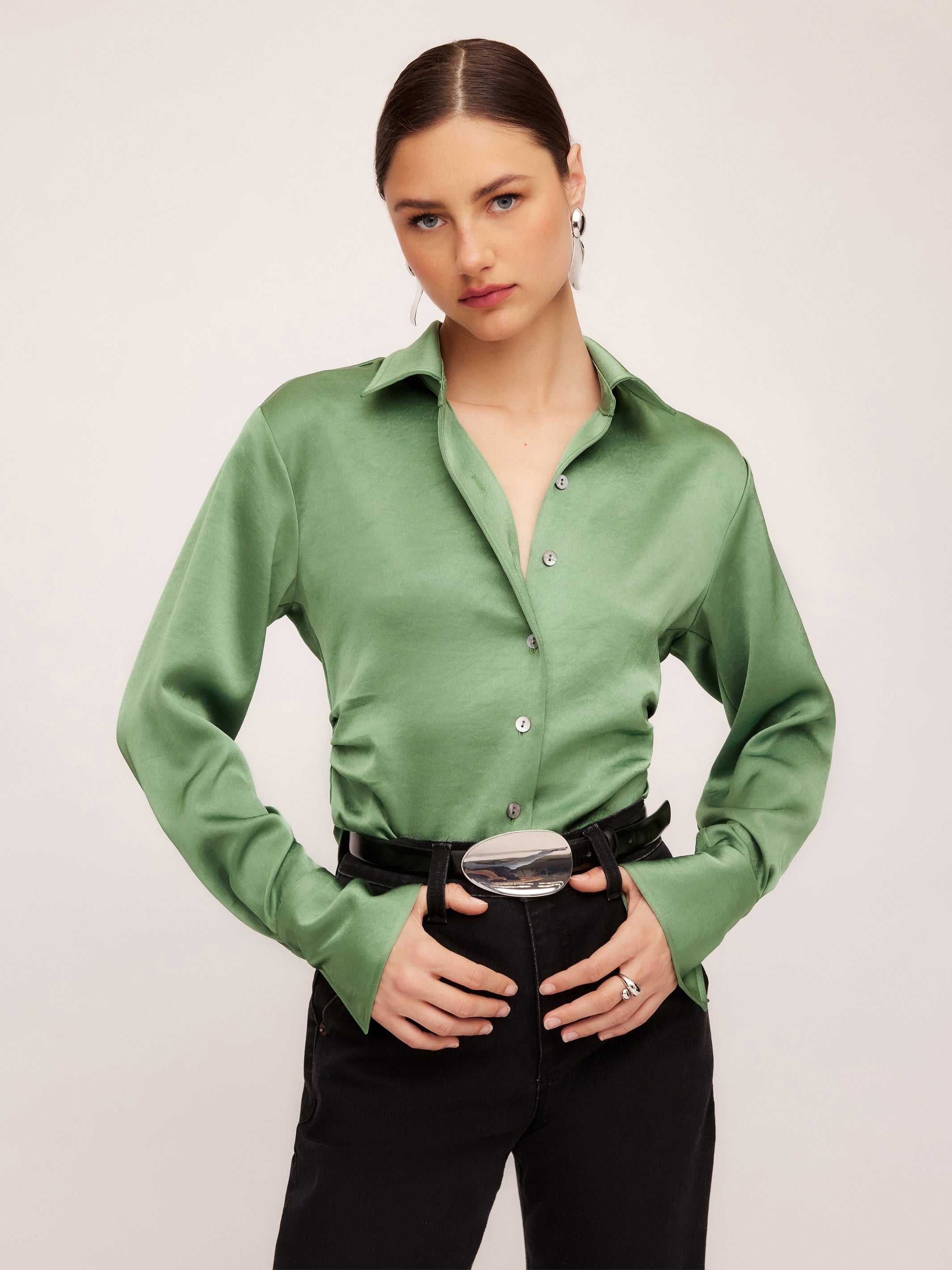 Woman wearing a green silky satin Tawnie Side-Pleat Shirt with cuffed long sleeves and button-down front tucked into black pants with a silver belt buckle.