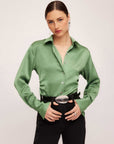 Woman wearing a green silky satin Tawnie Side-Pleat Shirt with cuffed long sleeves and button-down front tucked into black pants with a silver belt buckle.