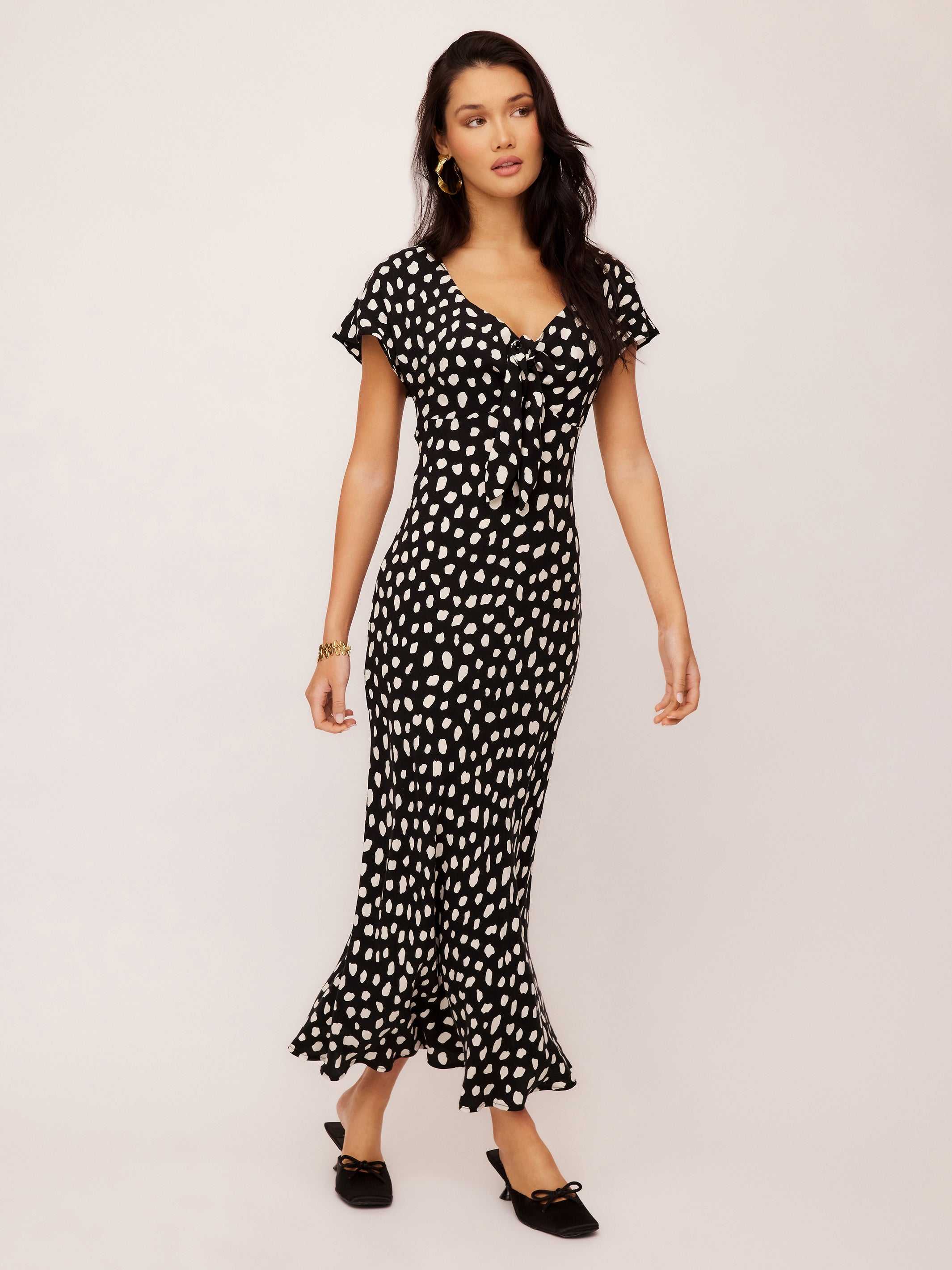 Woman wearing Emmeline Tie-Front black polka dot bias dress with short cape sleeves and ruffled hem