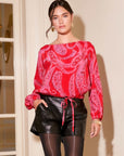 Woman wearing a scarlet paisley Claudine Tie-Waist Top with long bishop sleeves and black leather shorts.
