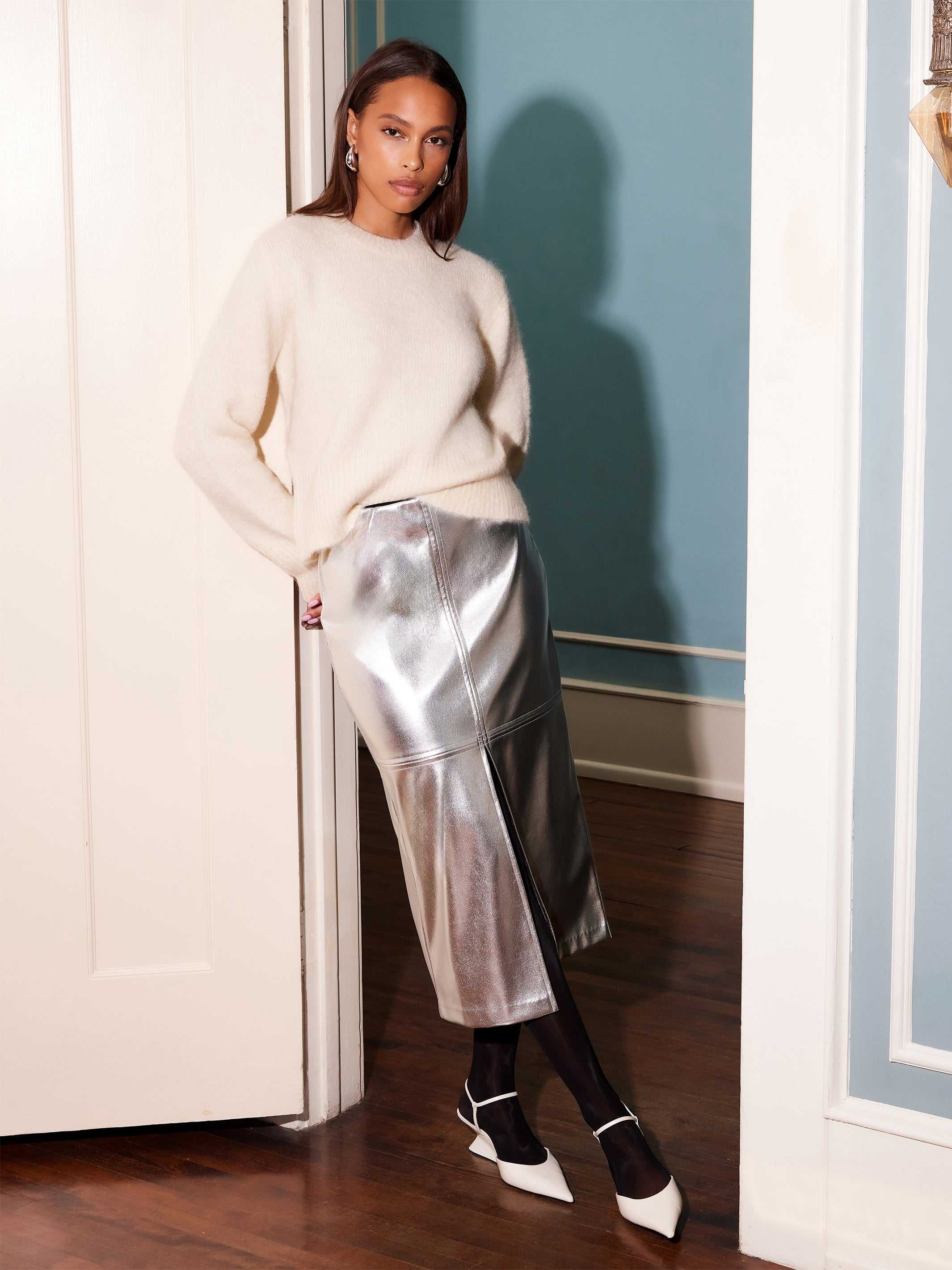 Woman wearing silver Kaia faux leather midi skirt with front slit and white sweater indoors