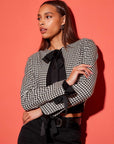 Woman wearing a cropped Poppy Bow Micro Jacket with herringbone check pattern, tie details at neck and sleeves, and long sleeves against a red background
