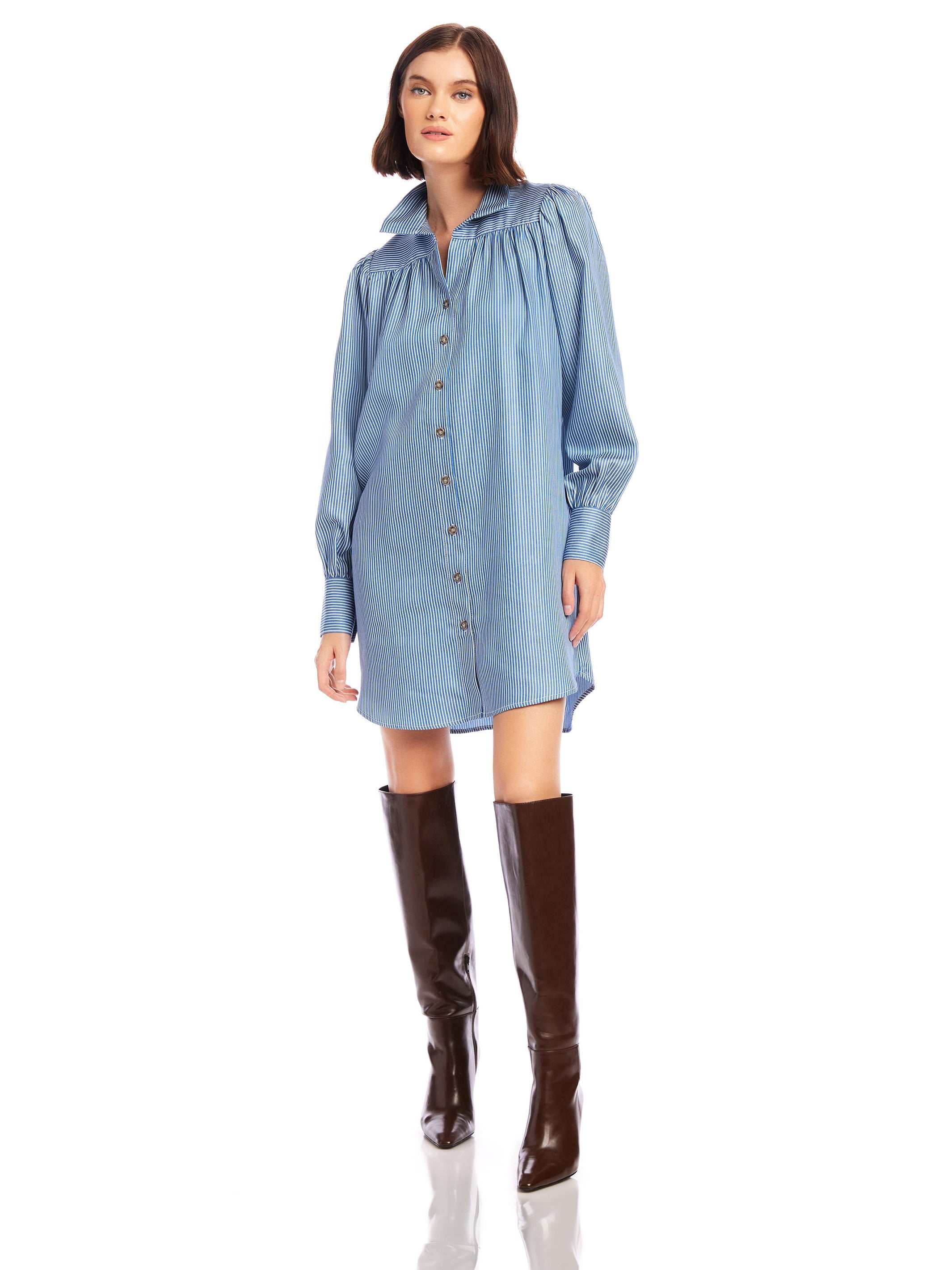 Woman wearing a light blue chambray stripe Natalia Shirtdress with long peasant sleeves and button front paired with knee-high brown boots.