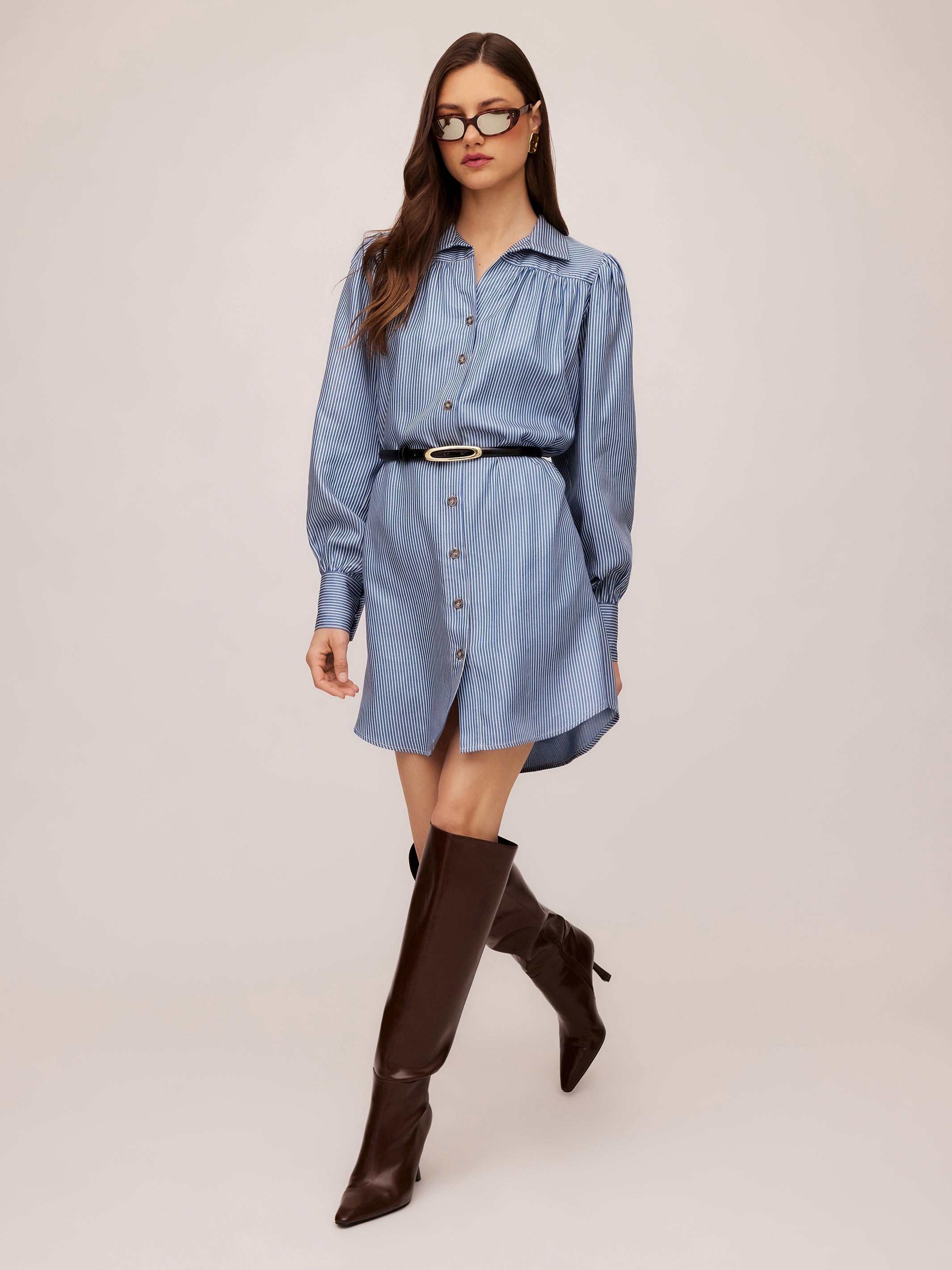 Woman wearing a blue Natalia Shirtdress with long peasant sleeves, button front, and black belt, styled with dark knee-high boots and sunglasses