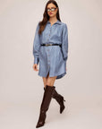 Woman wearing a blue Natalia Shirtdress with long peasant sleeves, button front, and black belt, styled with dark knee-high boots and sunglasses