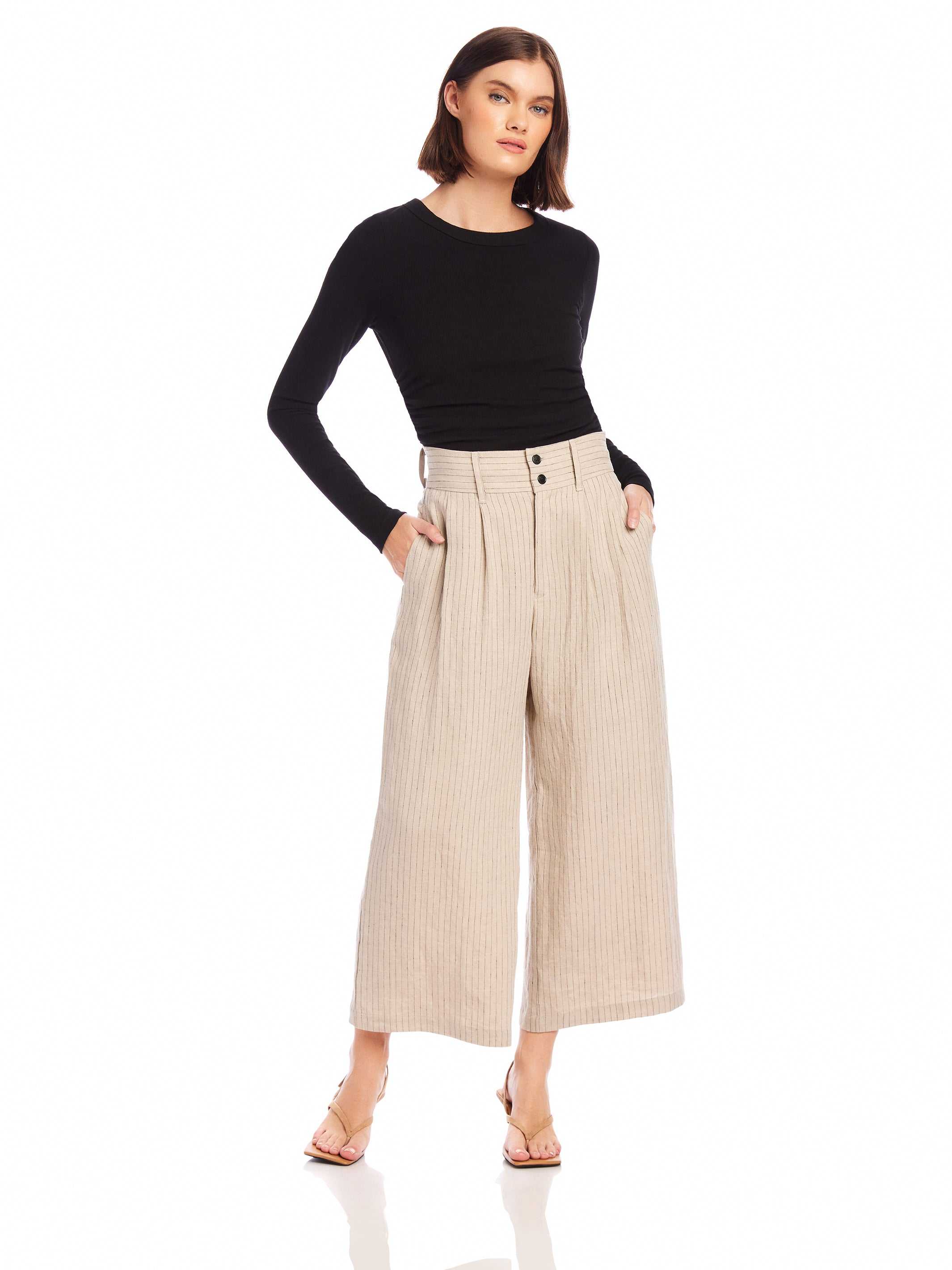 Woman wearing beige wide-leg cropped Sadie Trousers made of pinstripe linen with front pleats and side pockets paired with a black long-sleeve top.