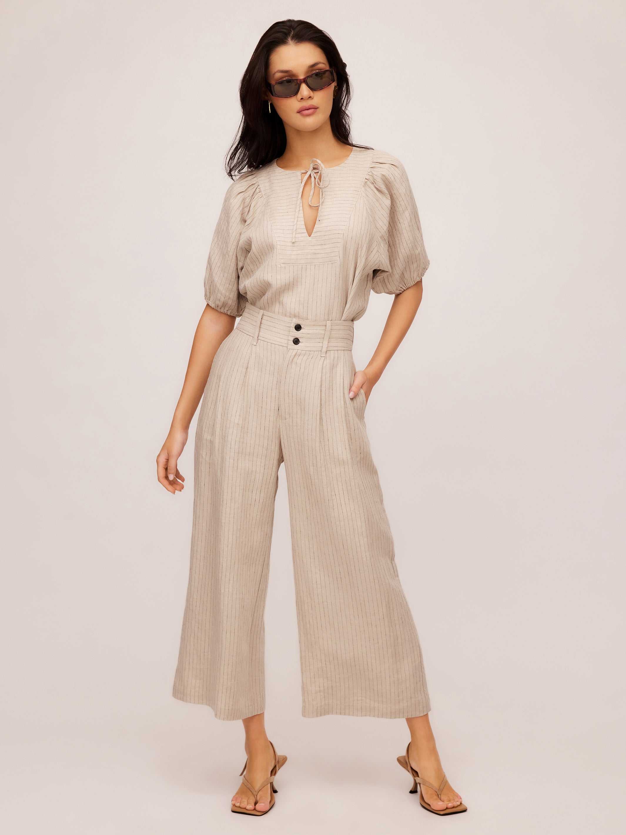 Woman wearing beige pinstripe linen Sadie Trousers with a wide leg and cropped length, styled with a light blouse and sunglasses.