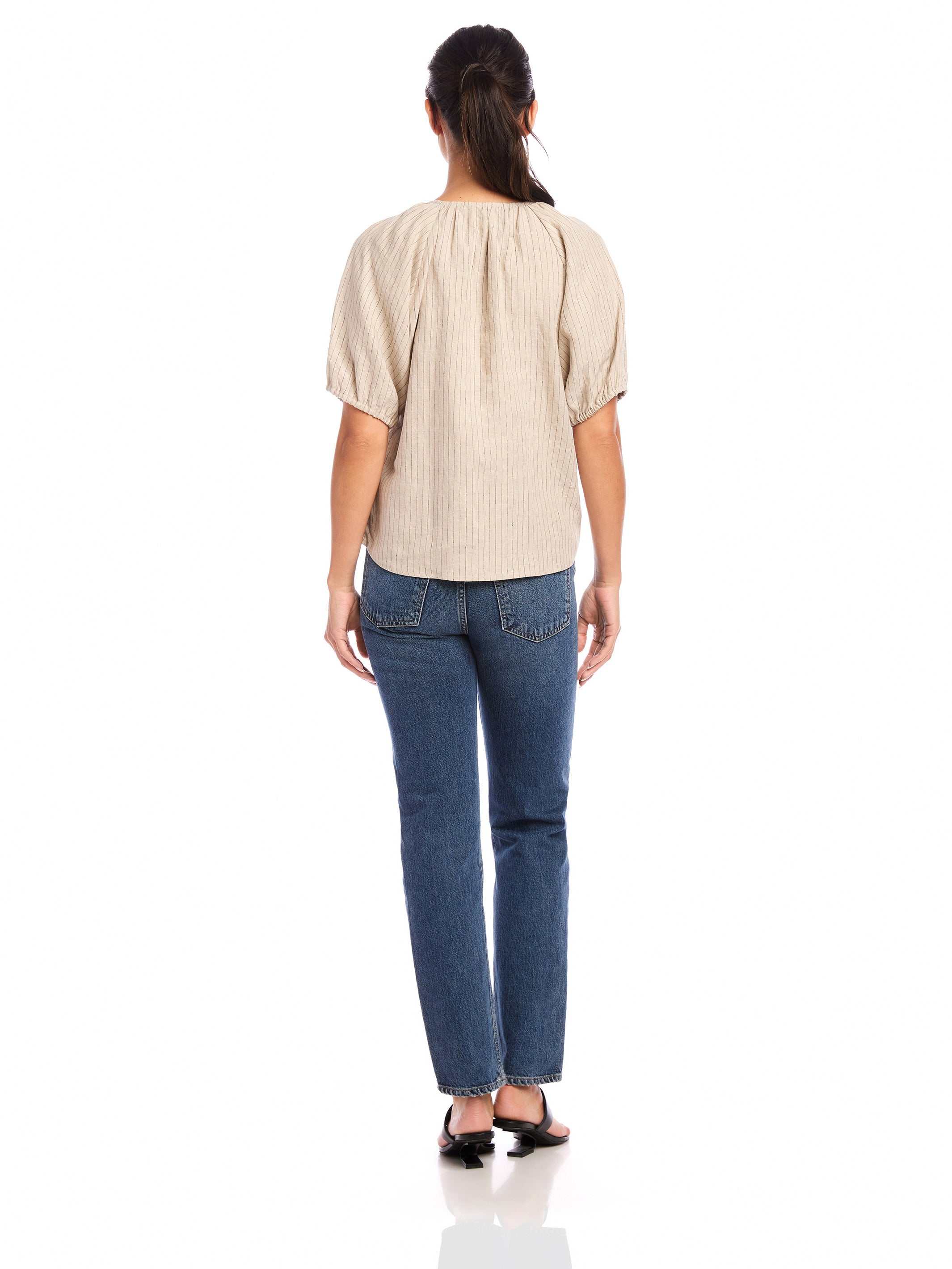Woman wearing beige pinstripe linen Imogen Poof Sleeve Top with short elasticized poof sleeves and split neck tie, paired with blue jeans, back view.