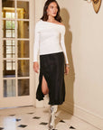 Woman wearing a black Brielle midi skirt with side slit and elastic waistband paired with white off-shoulder top and silver boots indoors