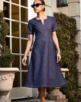 Woman wearing Olivia Contrast Stitch Dress in indigo twill with short sleeves and split neck standing outdoors near a door and greenery