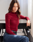 Woman wearing brick red Ruched Selena Top with ruched front, mock neckline, and long sleeves sitting on black chair