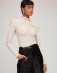 Woman wearing beige Ruched Selena Top with mock neckline and ruched front, paired with black sequined pants