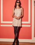 Woman wearing beige Safia Faux Suede Dress with spaghetti straps and square neck, paired with black tights and heels, standing against coral wall background