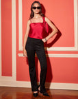 Woman wearing black Neli Slim Bootcut Pants with a red sleeveless top and black heels, posing indoors against a coral wall.