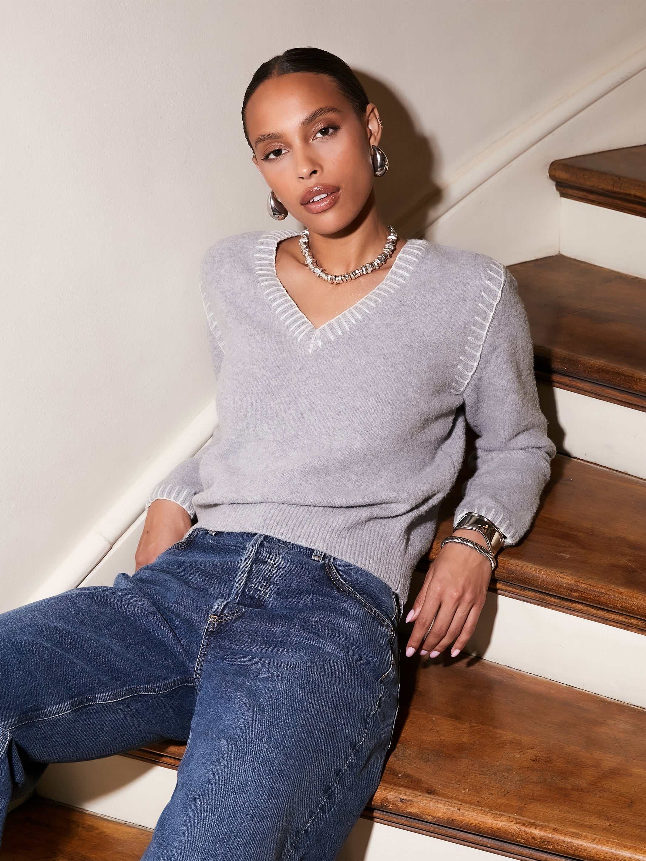 Woman wearing grey V-neck Ali Edge Stitch Sweater with white edge stitch detail and ribbed cuffs sitting on wooden stairs