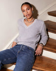 Woman wearing grey V-neck Ali Edge Stitch Sweater with white edge stitch detail and ribbed cuffs sitting on wooden stairs