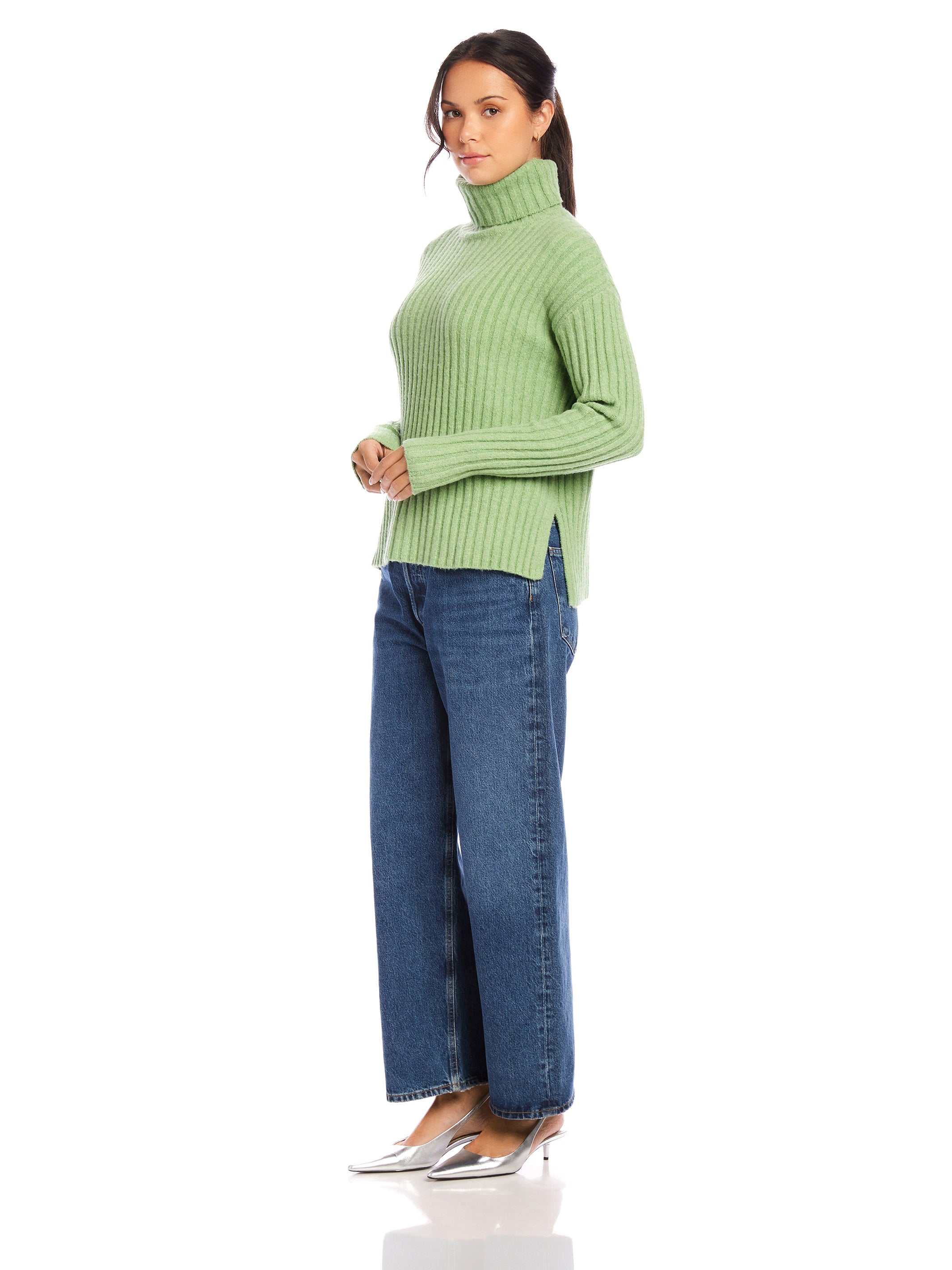 Woman wearing a sage green Claudia turtleneck sweater with wide rib detail and side slits paired with blue jeans and silver heels.