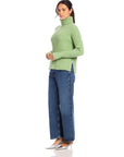 Woman wearing a sage green Claudia turtleneck sweater with wide rib detail and side slits paired with blue jeans and silver heels.