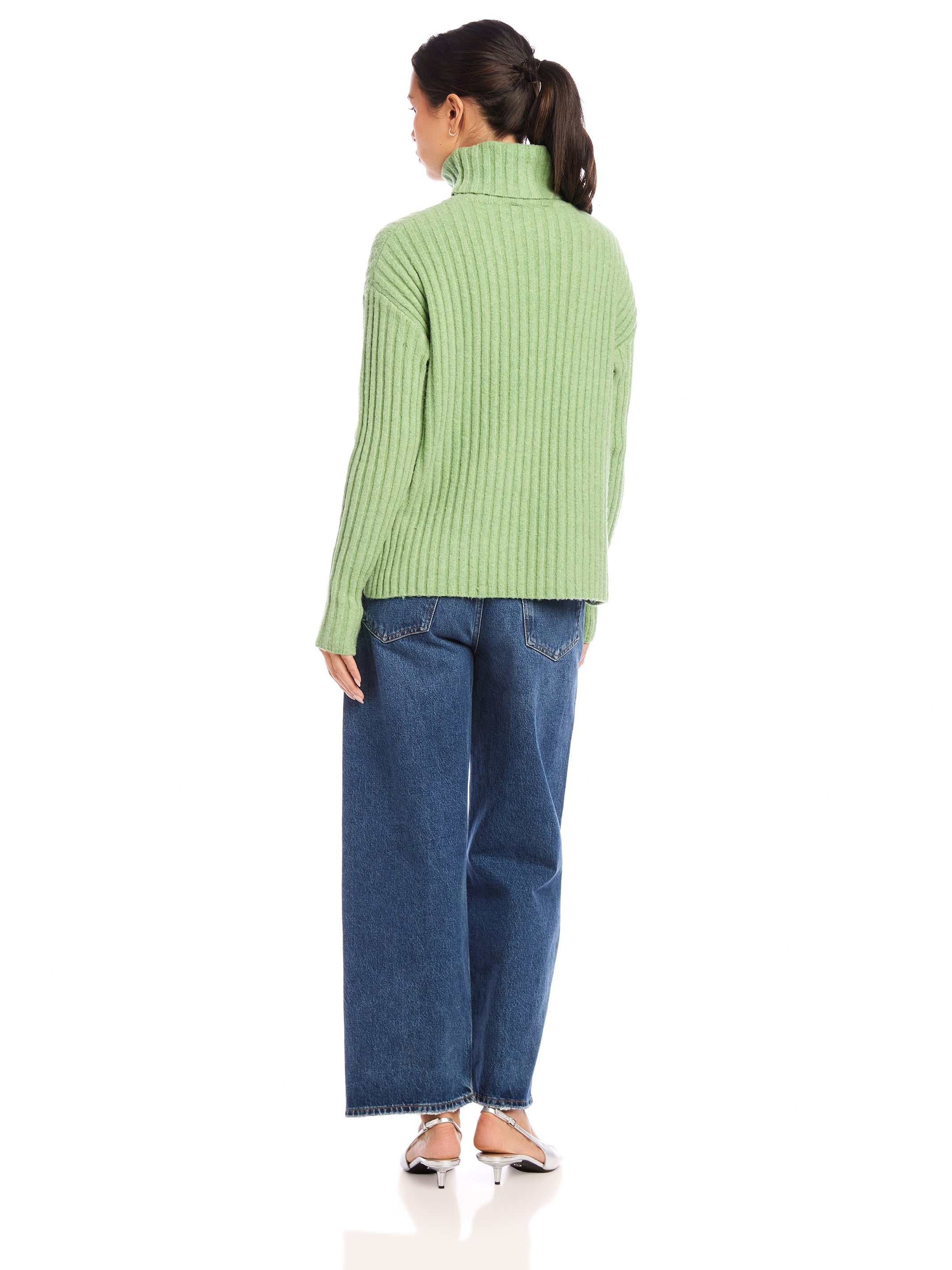 Back view of woman wearing sage green Claudia turtleneck sweater with wide rib detail and side slits, paired with blue jeans.