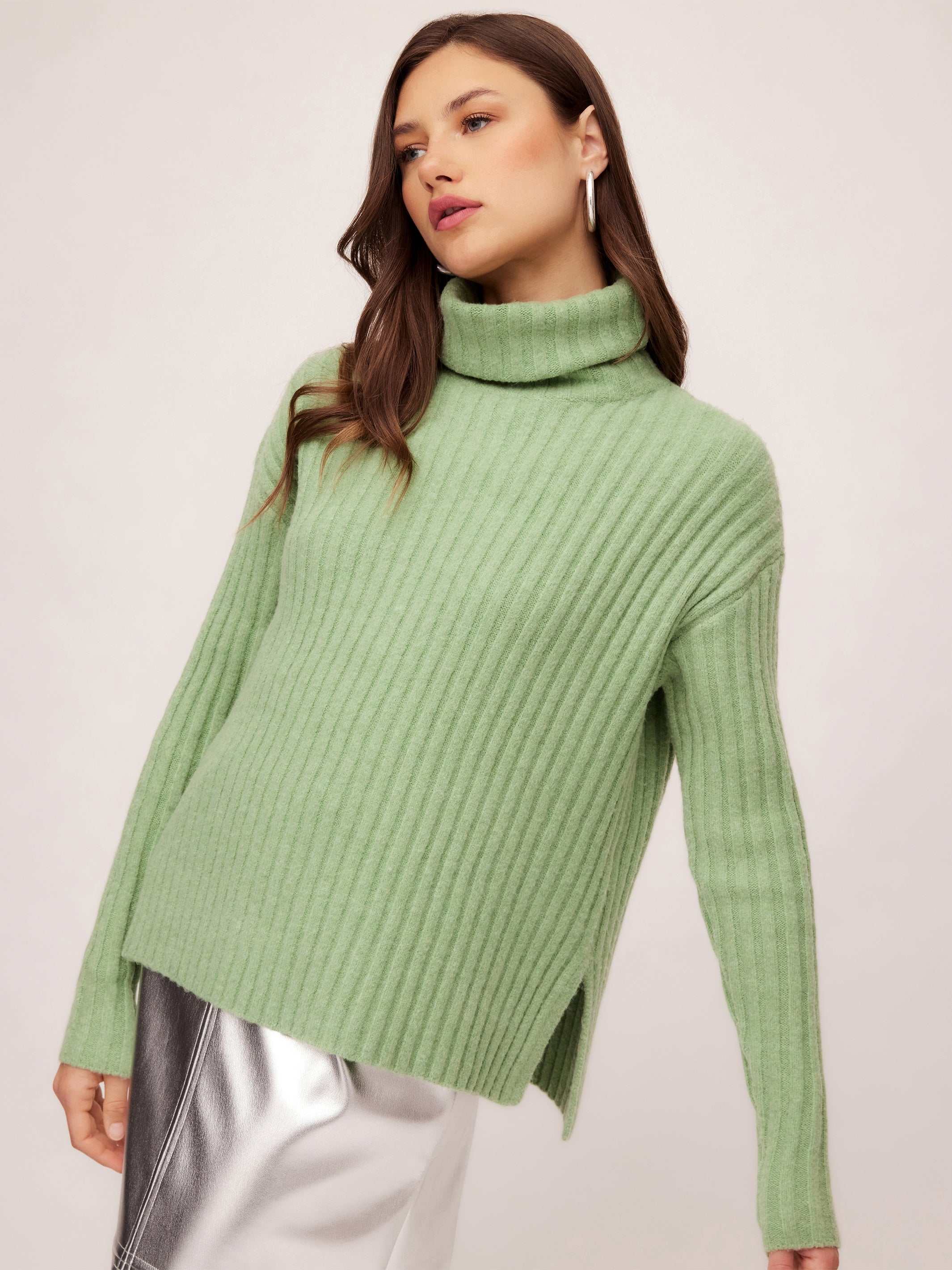 Woman wearing a sage Claudia turtleneck sweater with wide rib detail and side slits.